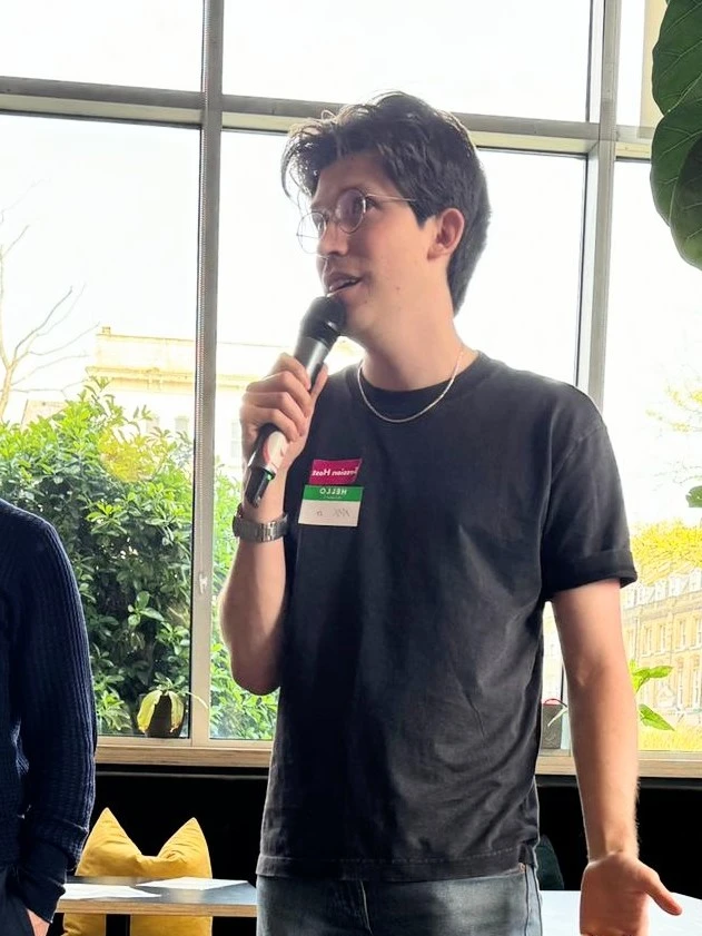 Max speaking into a handheld microphone at an event, wearing a session host badge.